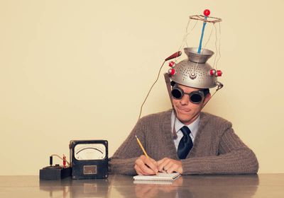 Conceptual retro image of a man wearing a silly mind reading gadget on his head, holding a pencil, and writing his thoughts down in a notebook.