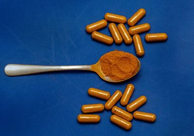 Orange powder in a silver spoon, surrounded by orange pills on a blue background.