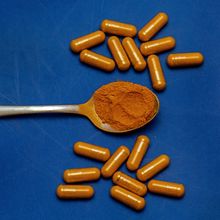 Orange powder in a silver spoon, surrounded by orange pills on a blue background.