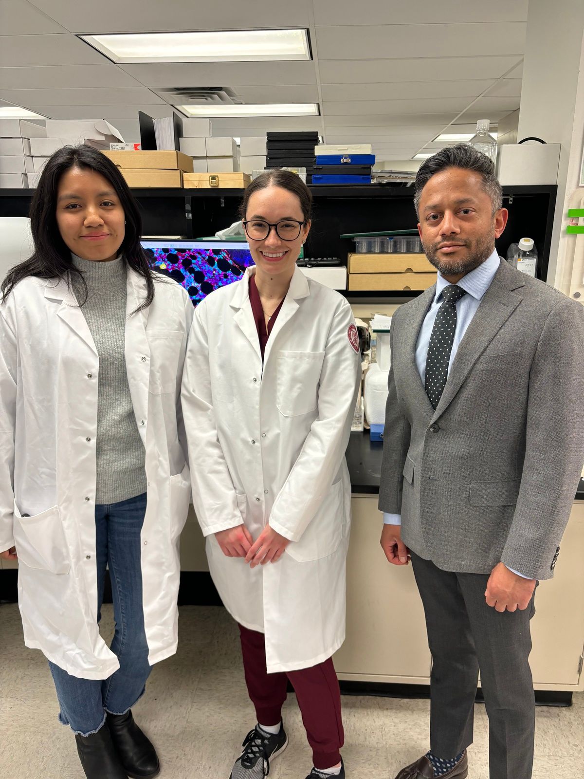 A photo of Weill Cornell Medicine scientists Itzel Valencia (left), Caitlin Unkenholz (middle), and Sanjay Patel (right) standing in front of a monitor that shows a tissue image produced by multiplex immunofluorescence. 