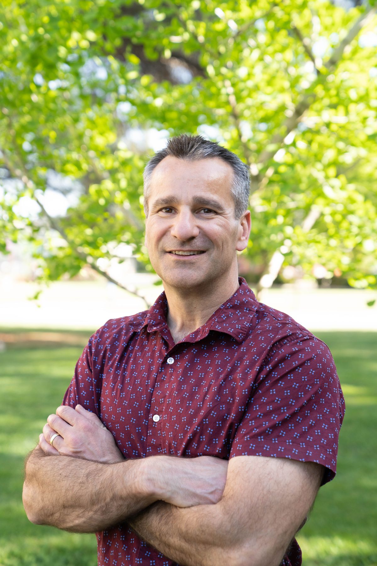 Keith Baar is wearing a patterned shirt. He is standing in front of a tree with his arms crossed.