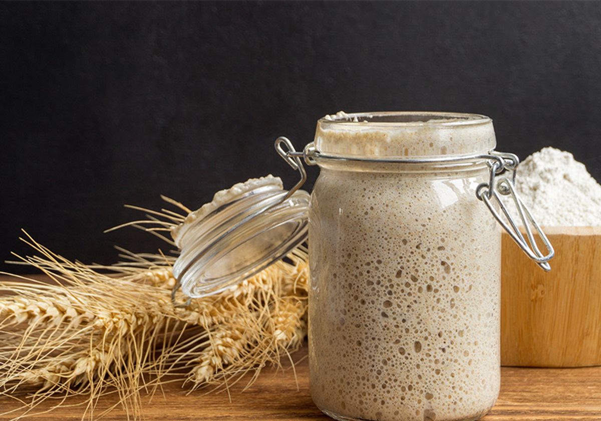 An active rye sourdough starter has risen to the top of a glass jar and sits in front of a bundle of dried rye plants and a container of flour.